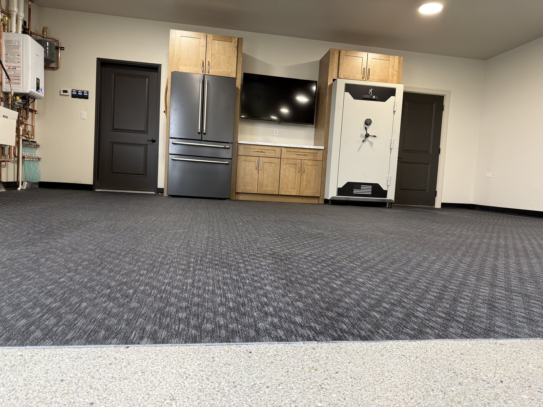 After: the lounge end of a Reiles Acres, ND garage with a wood-tone cabinet wall framing the refrigerator, TV and safe over rubber gym flooring with black vinyl base and a flake epoxy edge