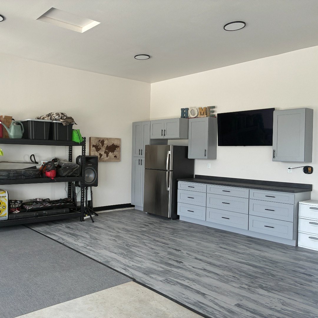 After photo of a Mapleton ND garage with a 16-foot gray shaker cabinet run, dark countertop, mounted TV and stainless refrigerator bay over new gray wood-look LVT flooring