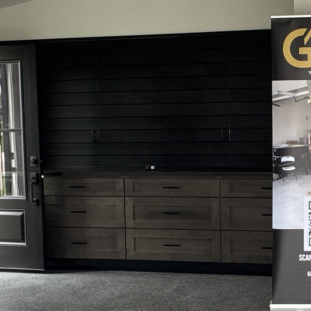 Close after photo of stained gray shaker drawer-base garage cabinets with black pulls, a dark laminate countertop and a black slat wall in a Mapleton ND garage