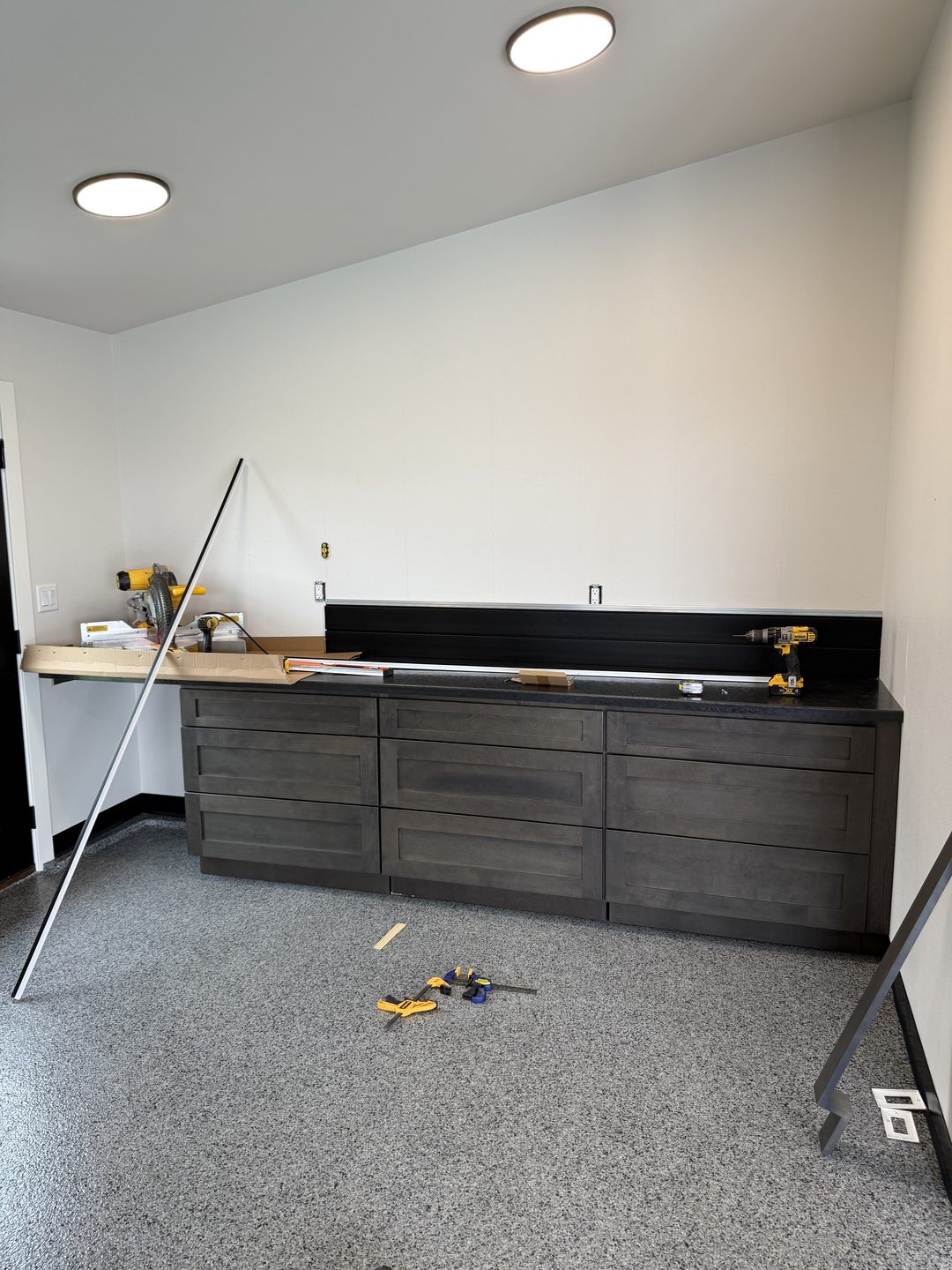 In-progress photo of stained gray shaker drawer-base garage cabinets with a dark laminate countertop and the first black slat wall panels in a Mapleton ND garage