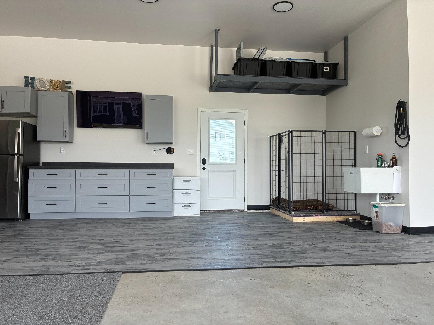 Wide after photo of a Mapleton ND garage bay showing the stainless refrigerator and garage cabinets on the left, the house entry door, and the dog kennel and utility wash sink on the right beneath a gray overhead storage rack