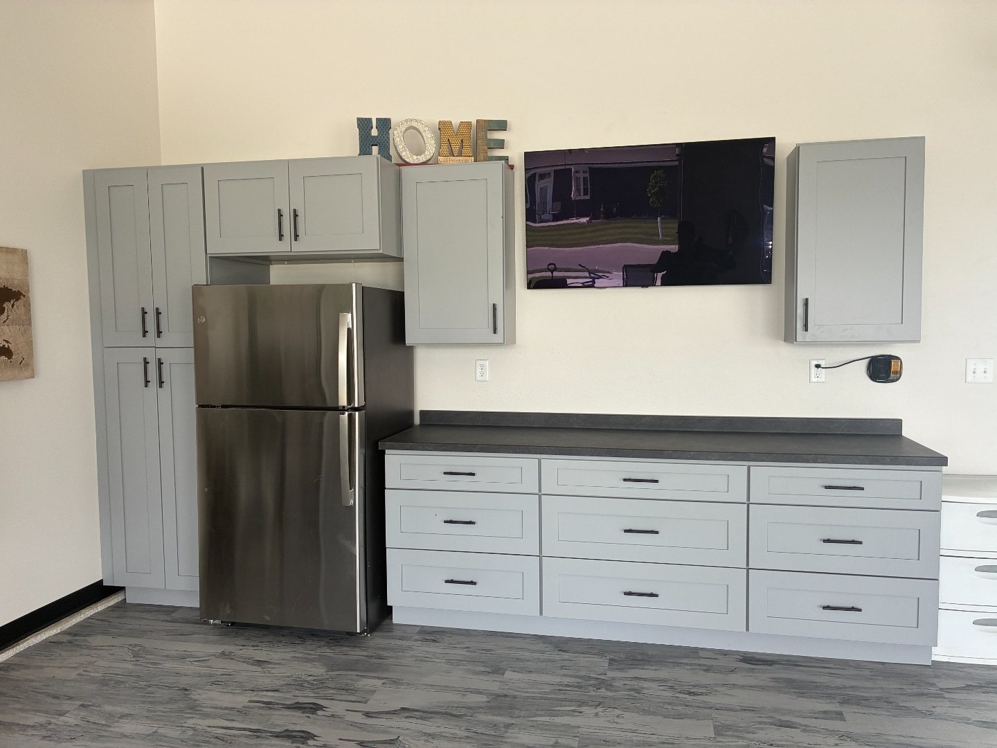 After photo of finished 16-foot gray shaker garage cabinets with dark countertop, wall uppers, mounted TV and stainless refrigerator bay in a Mapleton ND garage