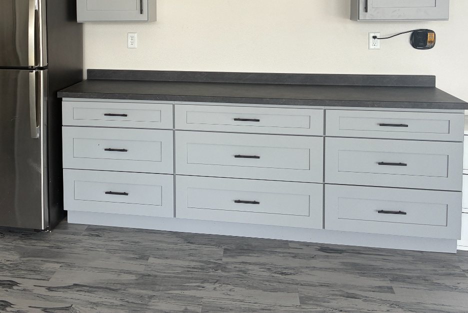 Close after photo of gray shaker garage base cabinets with soft-close drawers and a dark countertop above gray wood-look LVT flooring in a Mapleton ND garage