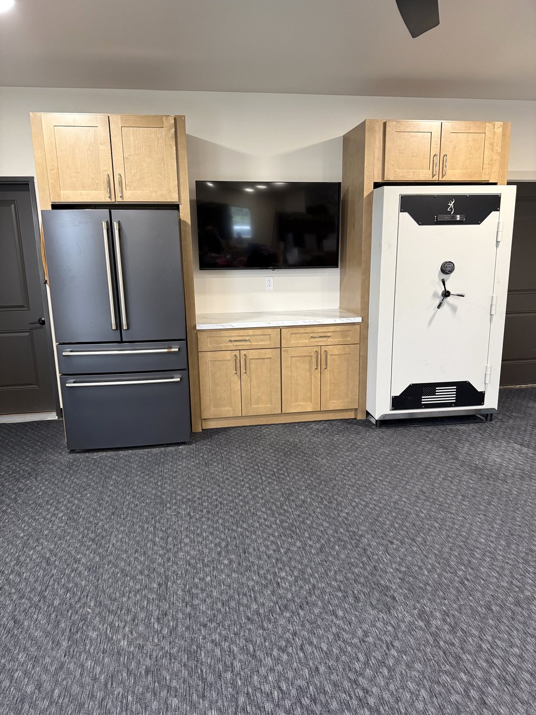 After photo of a natural wood-tone shaker cabinet wall with uppers framing a black refrigerator, a wall-mounted TV over a white laminate countertop, and a safe, above rubber gym flooring in a Reiles Acres ND garage