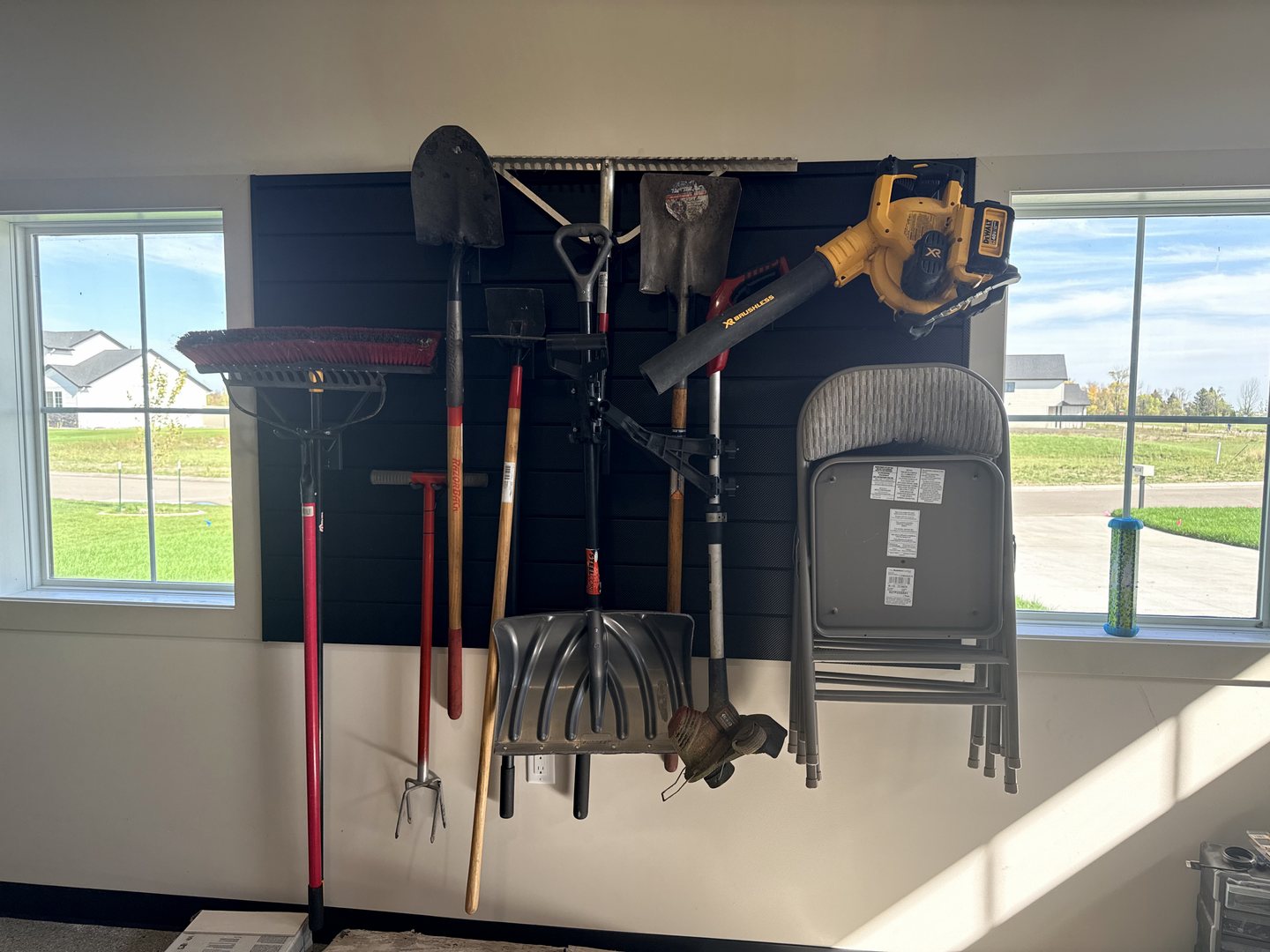 After photo of a black slat wall between two windows holding shovels, rakes, a leaf blower and folding chairs in a Reiles Acres ND garage